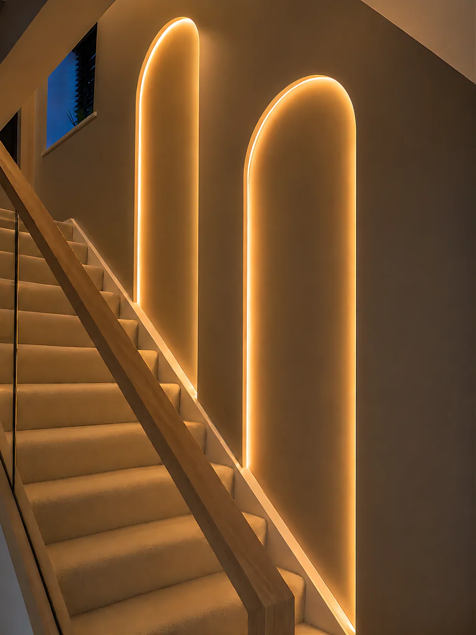 Two tall arches traced in continuous light on the wall beside a carpeted staircase, the timber handrail and treads washed by a low line of light.